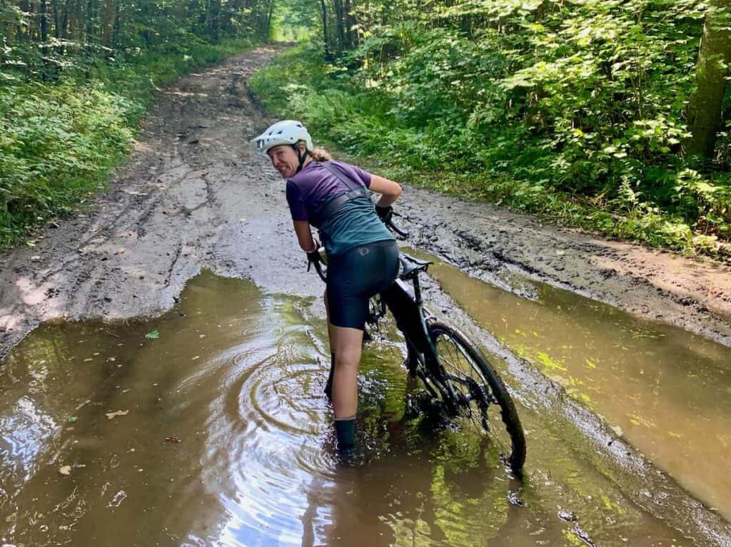 Woman standing in big mud puddle next to bike with grimace on face after bike stopped in the middle